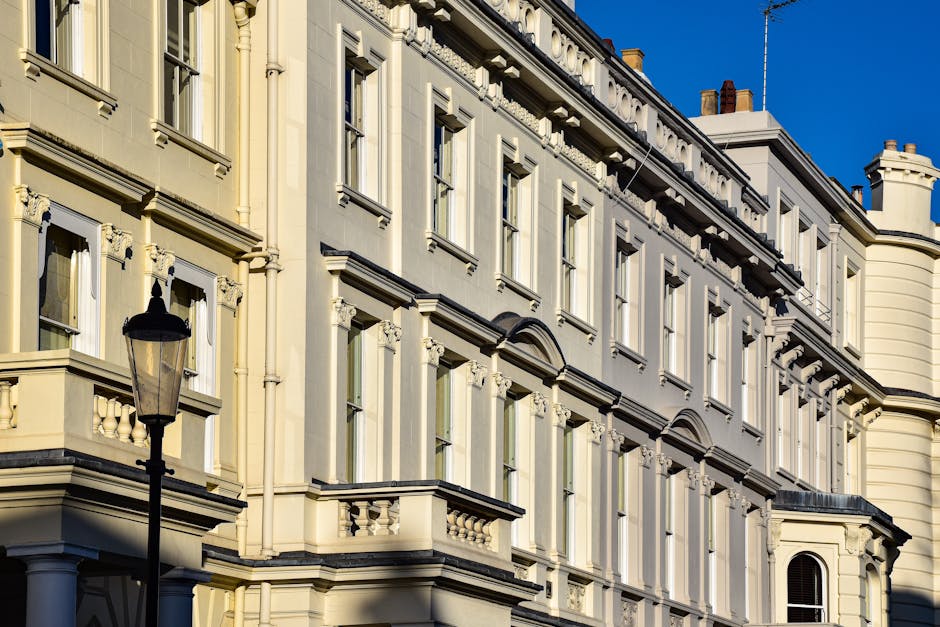 Photograph of a multi-story residential building with a classical architectural style, featuring cream-colored walls, decorative window frames, and ornate cornices. Several large windows with white tr