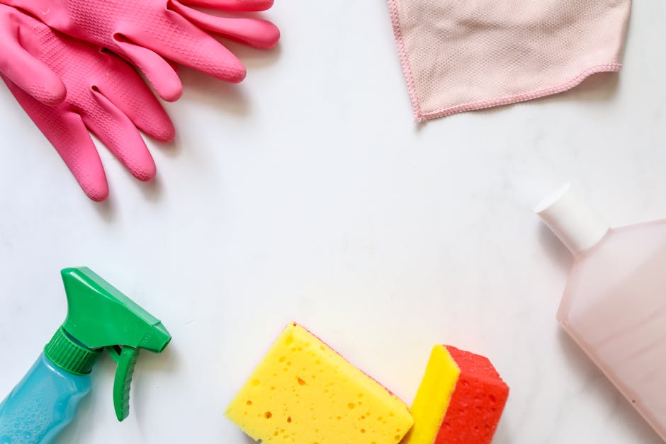 A flat lay of cleaning equipment arranged on a white surface, including a pair of pink rubber gloves at the top left, a pink cloth at the top right, a green spray bottle with a blue body at the bottom