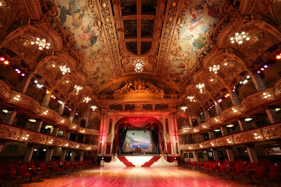 The image depicts the ornate interior of the Olympia London venue, featuring a large, polished wooden dance floor at the center with a red and white carpeted staircase leading to a stage. Surrounding the stage are intricate gold-colored architectural details on the balconies and walls, adorned with elaborate moldings and decorative panels. The high ceiling showcases painted frescoes and decorative plasterwork, with multiple elegant chandeliers providing soft lighting. The balconies are lined with red velvet chairs, and the overall space appears clean and well-maintained, suitable for hosting hire events. The lighting highlights the grandeur of the venue’s classical design, emphasizing its pristine condition and suitable environment for cleaning and sanitisation processes, which are managed by professionals like Cleaners SW5.