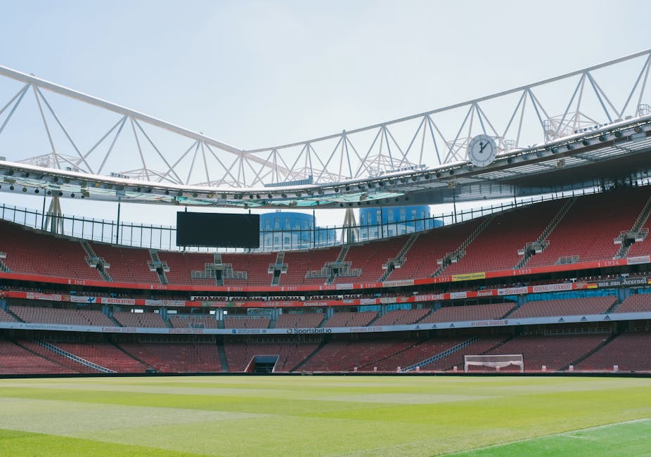 A view of an empty outdoor stadium with a vibrant green football pitch at the center, surrounded by red seating areas. Overhead, the stadium's retractable roof features a beige fabric covering supported by a network of steel cables and beams, with a portion of the blue sky and scattered clouds visible through the open sections. The stadium appears well-maintained and clean, suitable for hosting large sports events. This image exemplifies the importance of thorough surface cleaning and maintenance for venue hygiene, consistent with the cleaning checklist for hire events at Olympia London as detailed by Cleaners SW5.
