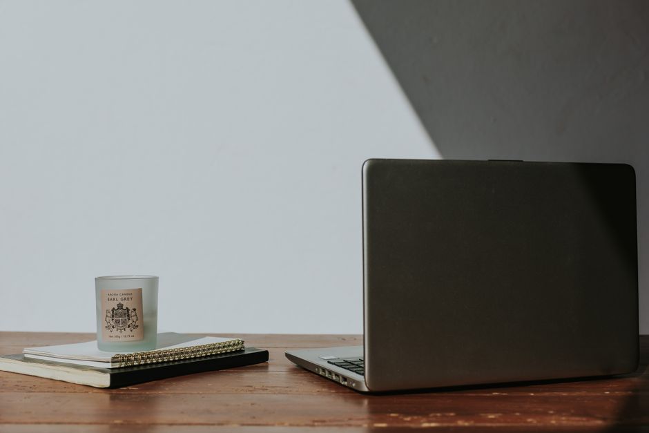 A close-up view of a wooden desk featuring a closed silver laptop on the right, with a small white candle in a glass container displaying a crest and the words 'Earl Grey' on the left. Next to the candle, there is a black notebook and a spiral-bound notepad. The desk surface appears clean and well-maintained, emphasizing surface cleaning and tidiness suitable for professional domestic or commercial cleaning services. The background includes a plain wall with soft lighting, highlighting the pristine condition and organized layout, reflecting thorough sanitisation and deep cleaning practices promoted by Cleaners SW5 near Earls Court Station, SW5.